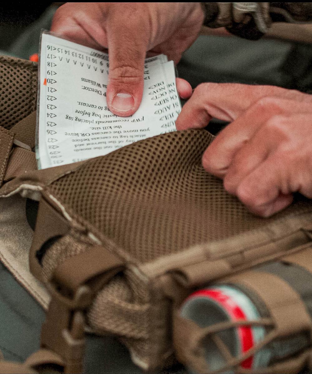Hands put white checklist into brown mesh tactical vest pocket.