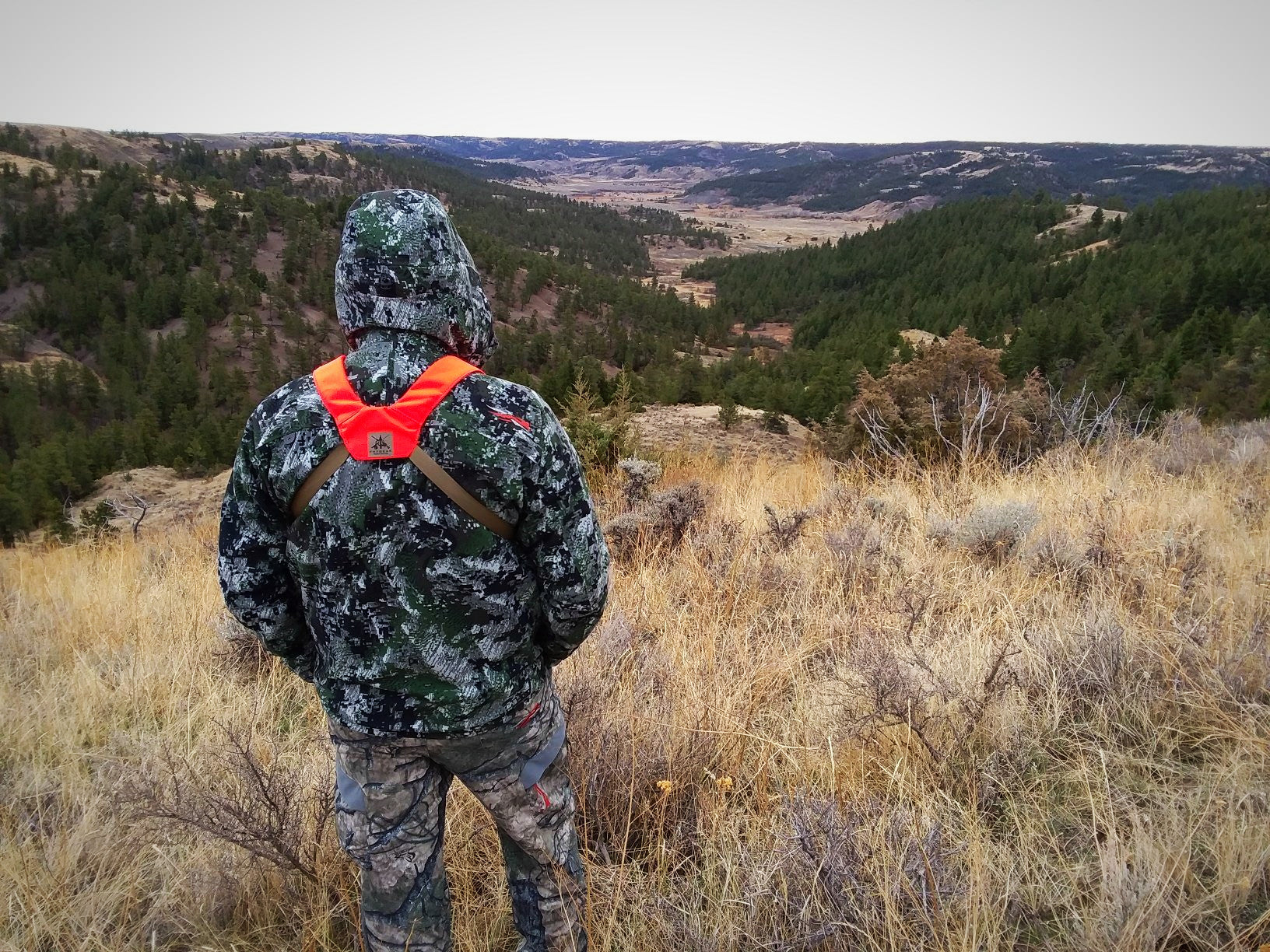 Hunter's hooded camo jacket & pants with orange chest harness overlooks a valley.