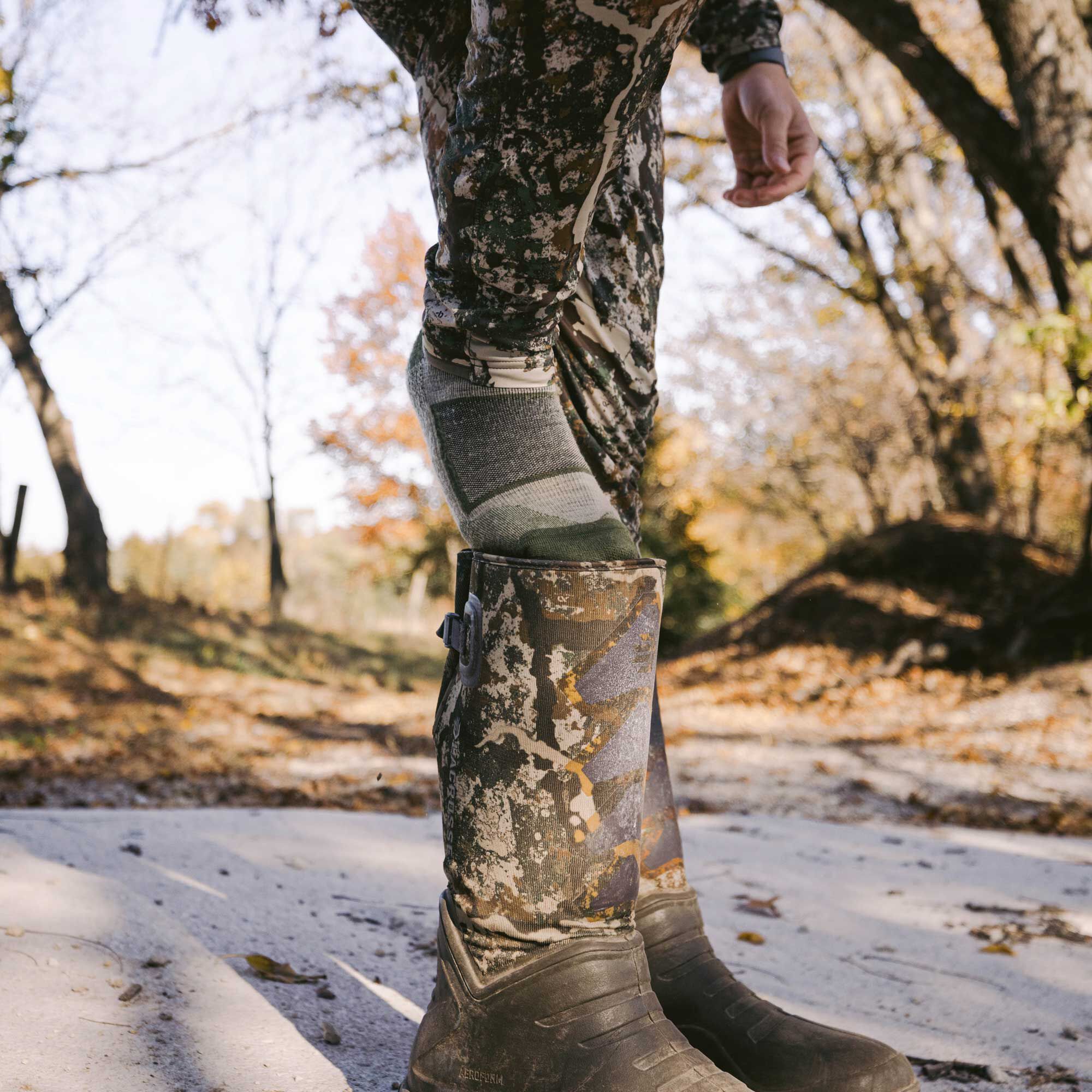 A grey and olive green patterned sock is pulled partially out of a muddy camouflage rubber boot worn with camouflage pants.