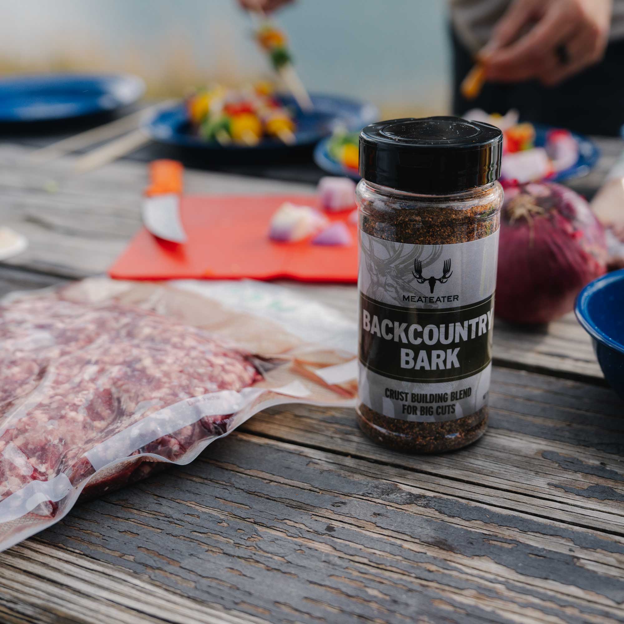 Clear jar of "Backcountry Bark" seasoning, black lid, on a wooden table.