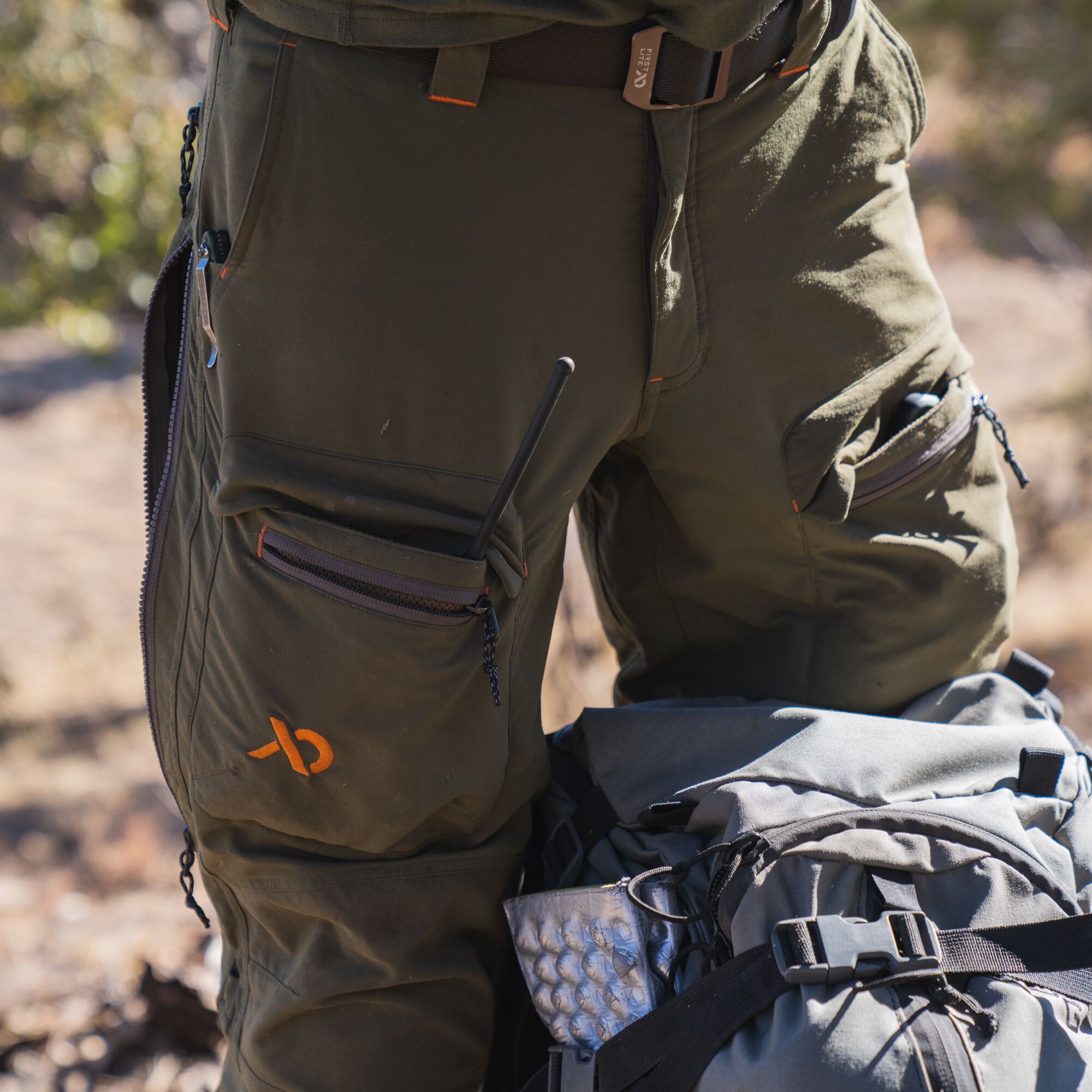 Dark olive green cargo pants with zippered pockets, an orange logo, orange stitching accents, and a side leg zipper.