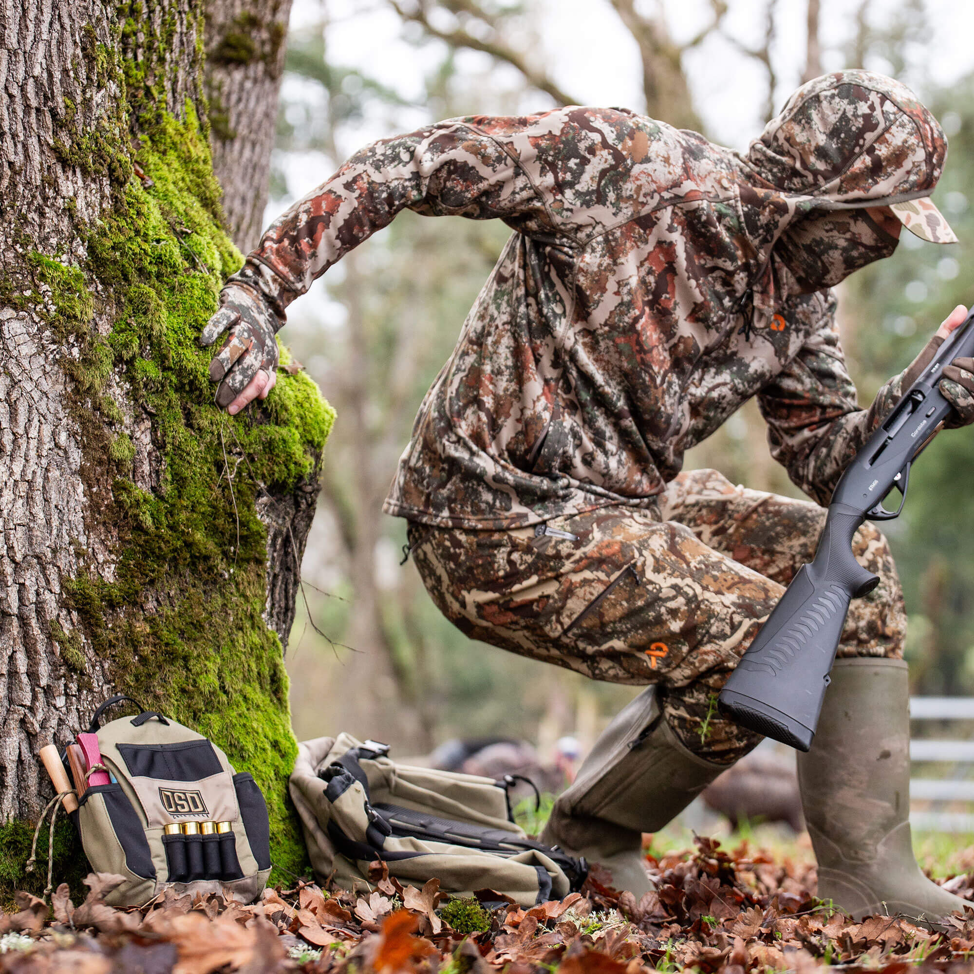 Hunter in multi-color camo suit holds shotgun, with gear bags by mossy tree.