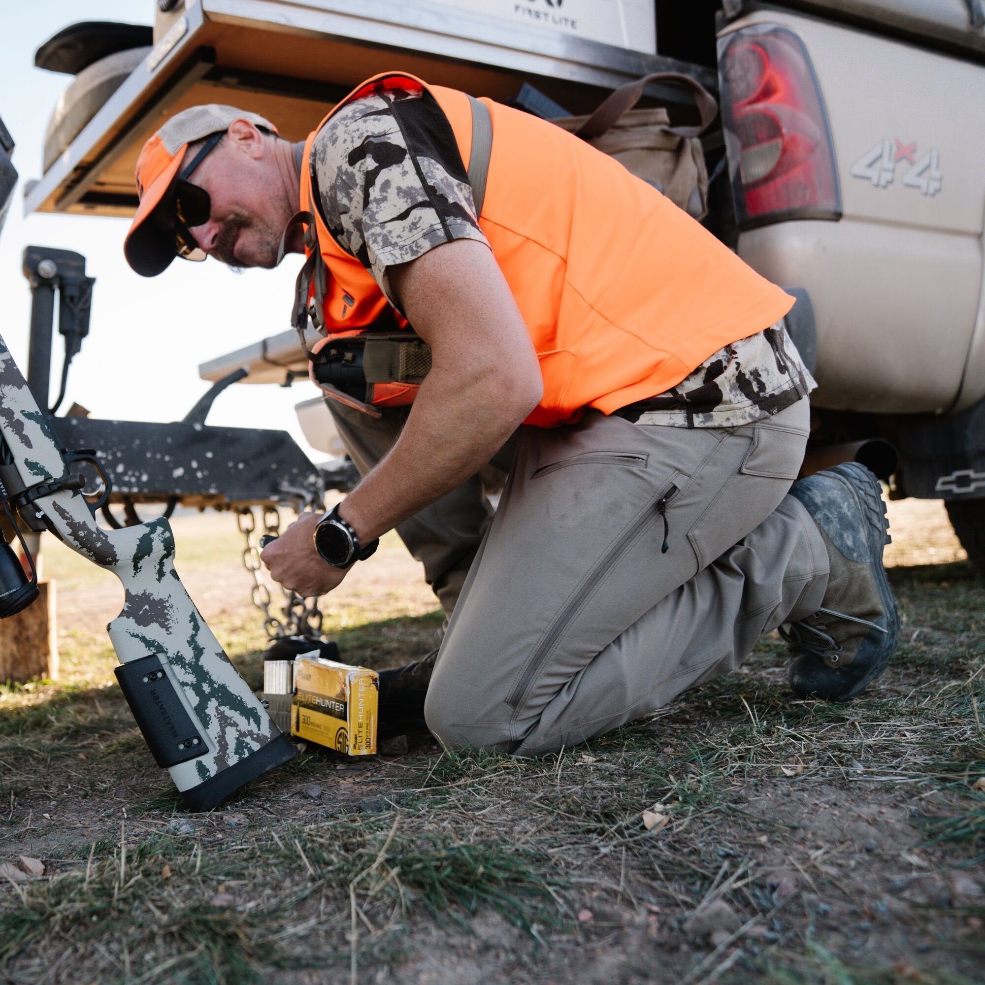 Man kneeling, wearing tan cargo hunting pants with zippered pockets, next to a camouflage rifle.