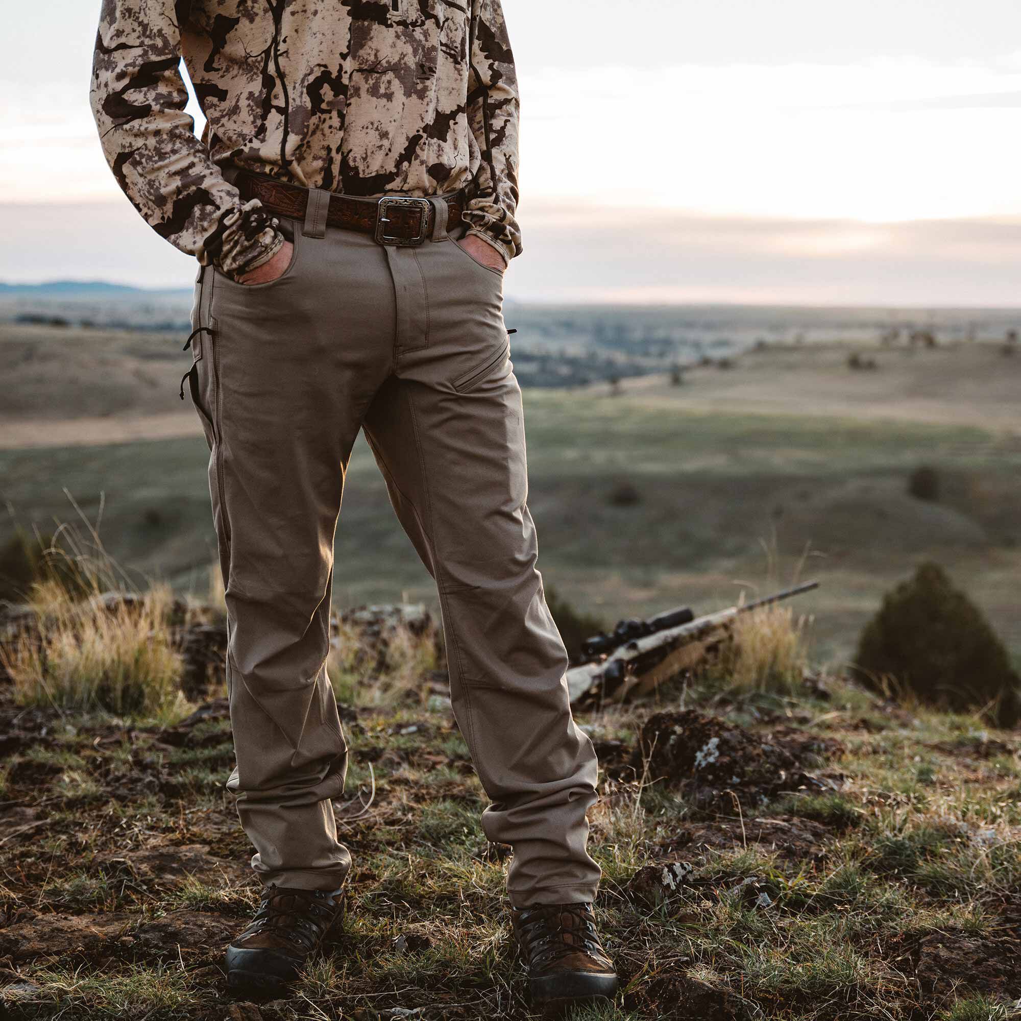 Men's khaki outdoor pants featuring articulated knees, zippered thigh pockets, and a brown leather belt, worn with a camo shirt and hiking boots.