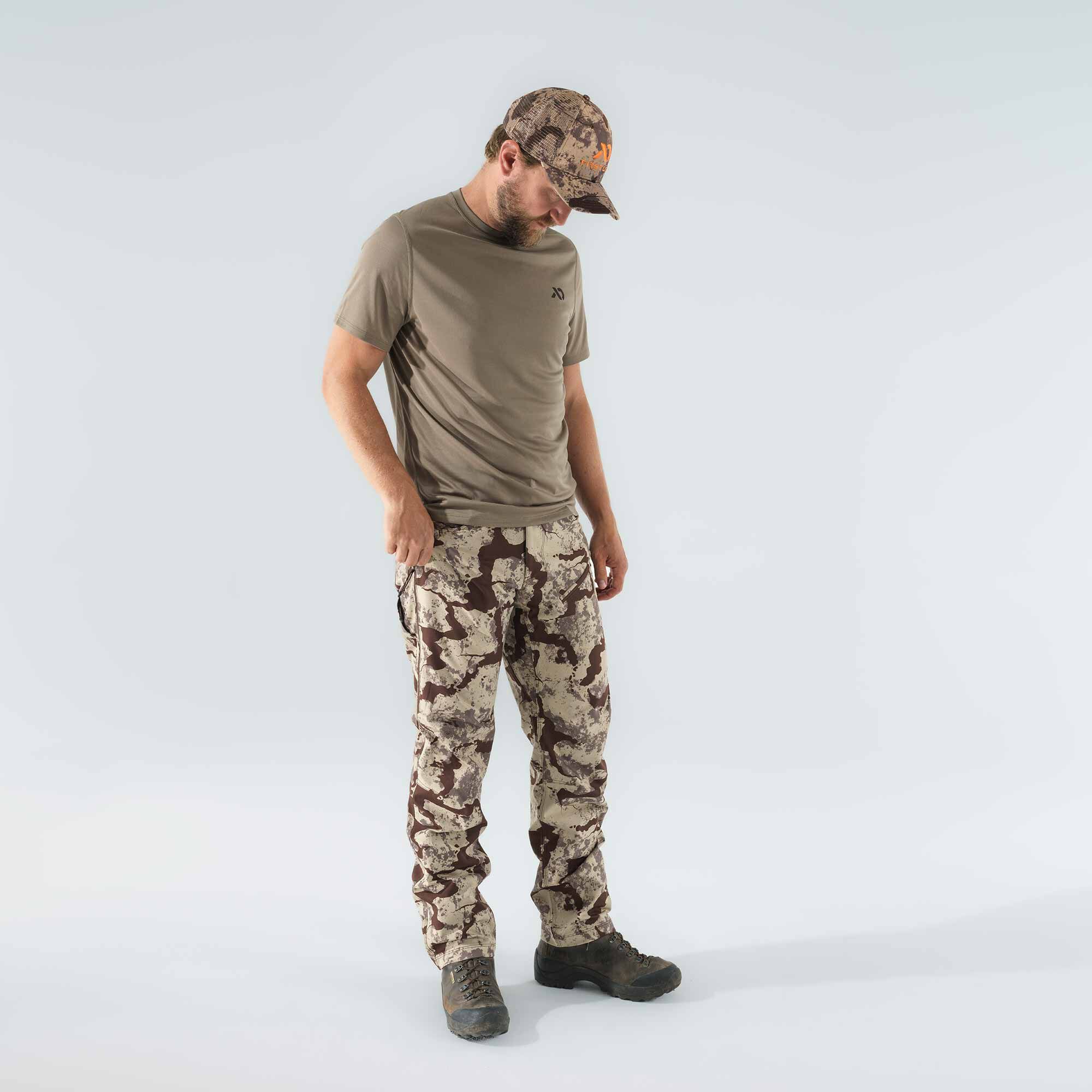 A man models an olive green t-shirt, brown and tan camouflage pants, and a matching camouflage cap.