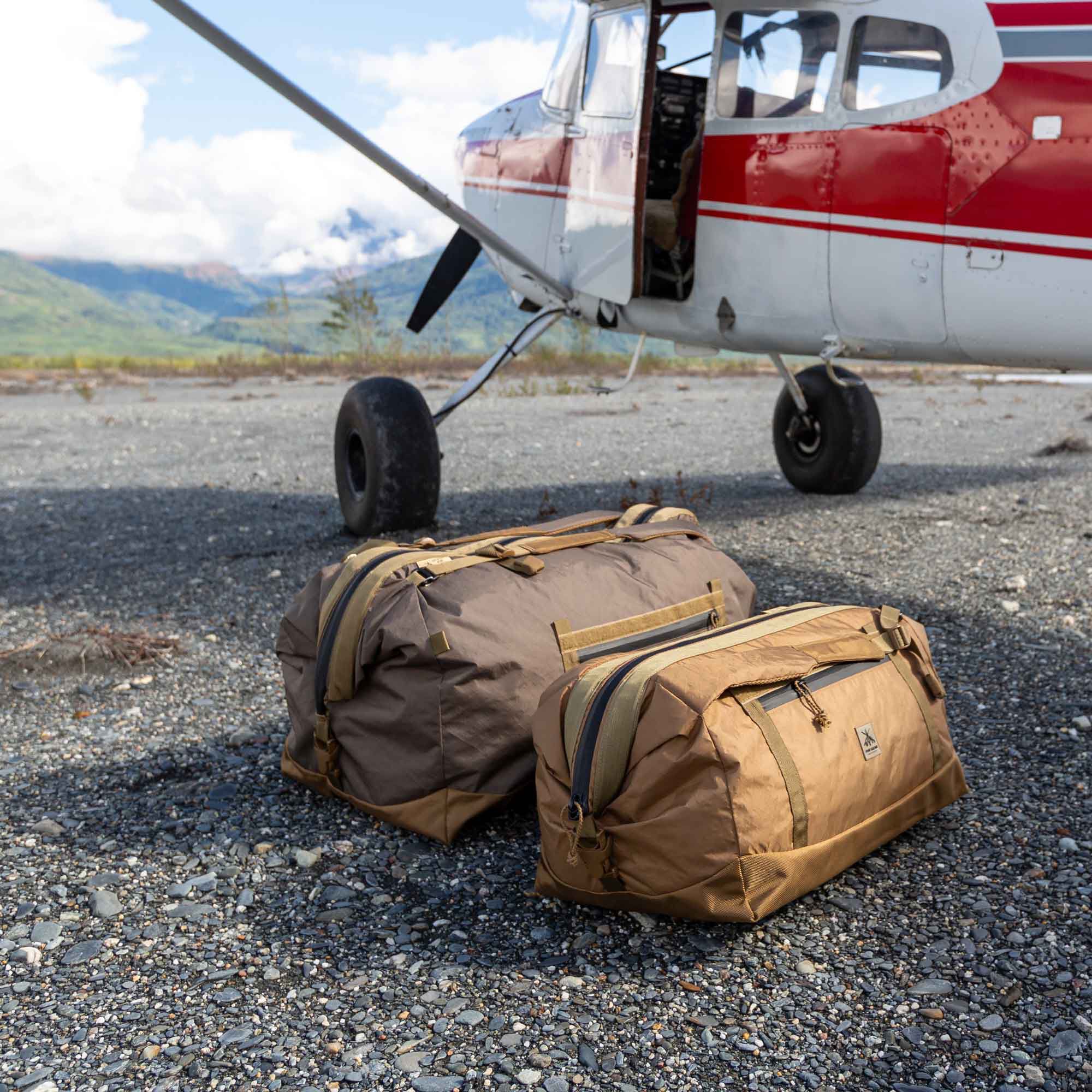 Two brown duffel bags with zippers and straps on gravel by a bush plane.
