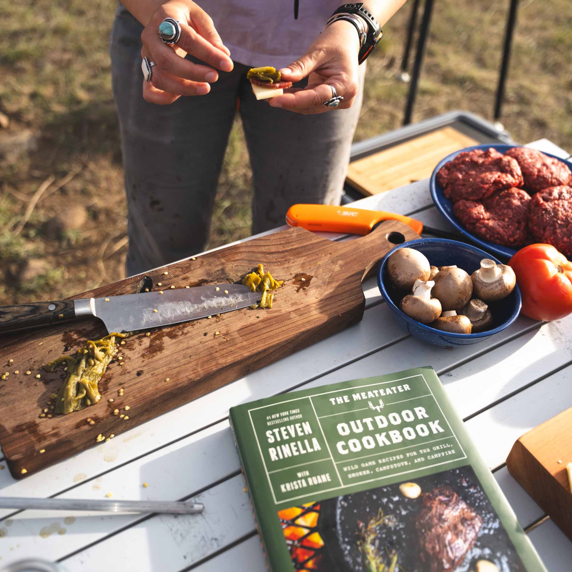 Person preparing food outdoors with a cutting board, knife, and 'The MeatEater Outdoor Cookbook' on a table.