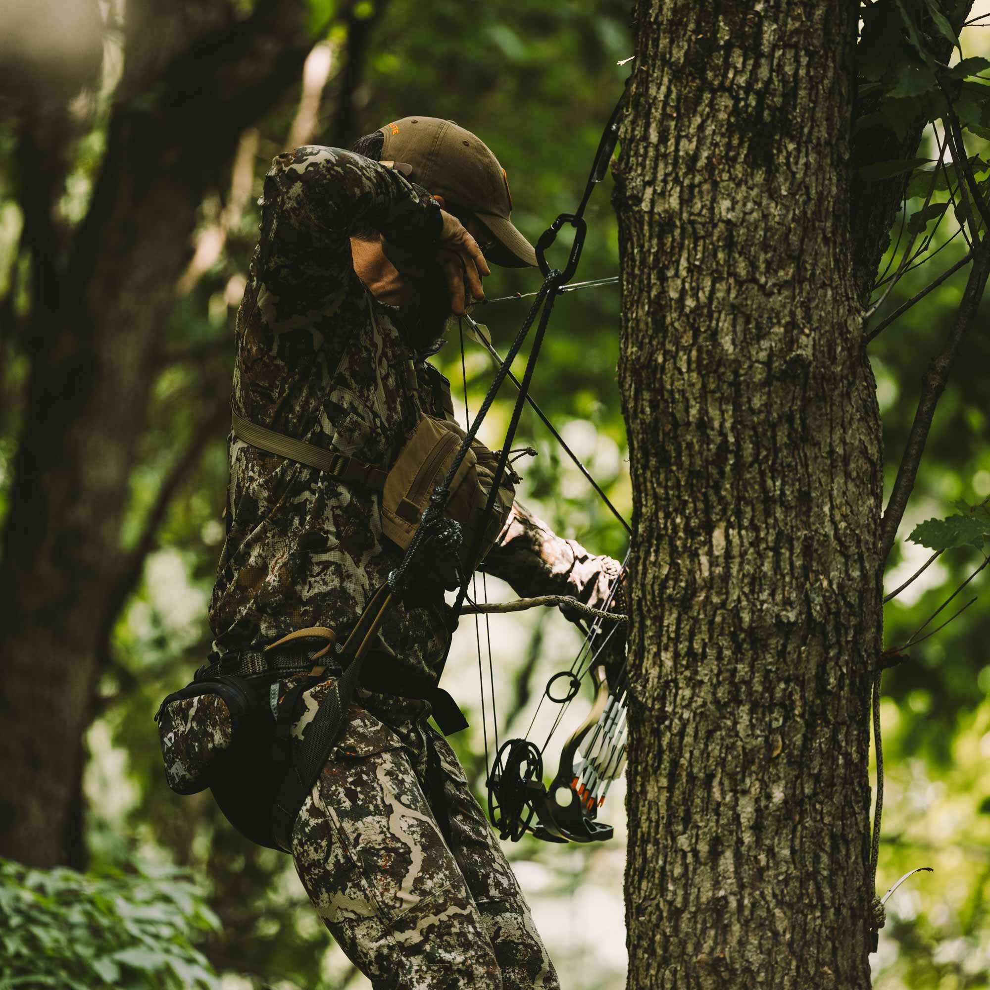 Camo hunting tree saddle on hunter, with bow, pouches, and tether to tree.