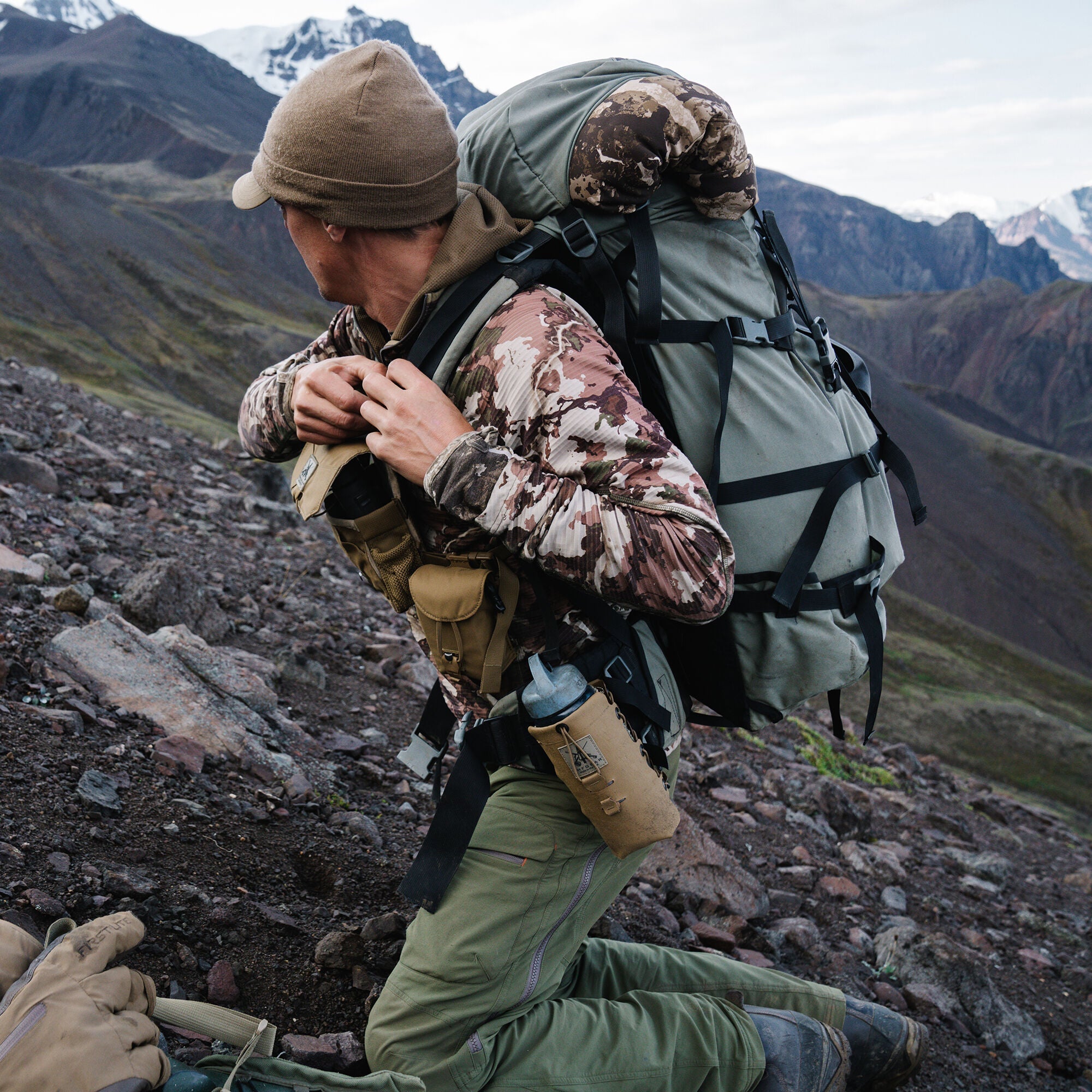 Camo top, green backpack, and pants with tan pouches worn by kneeling person outdoors.