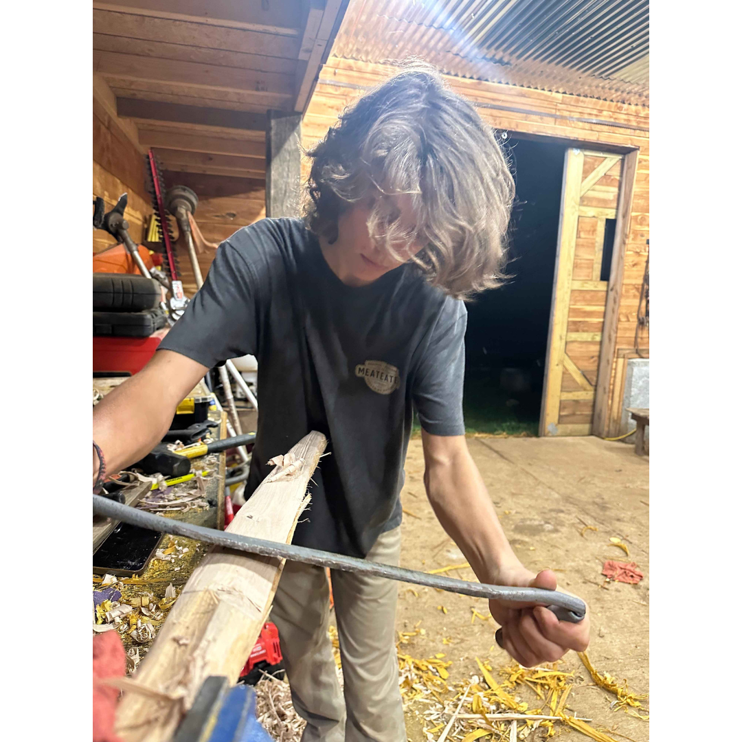 Young person in dark gray MEATEATER tee shaping light wood with a metal tool.