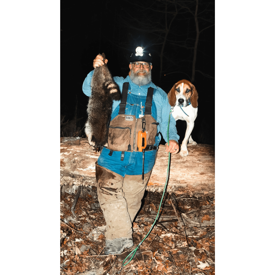 Man wears tan Dan's protective hunting gear with orange device, holds raccoon, dog nearby.