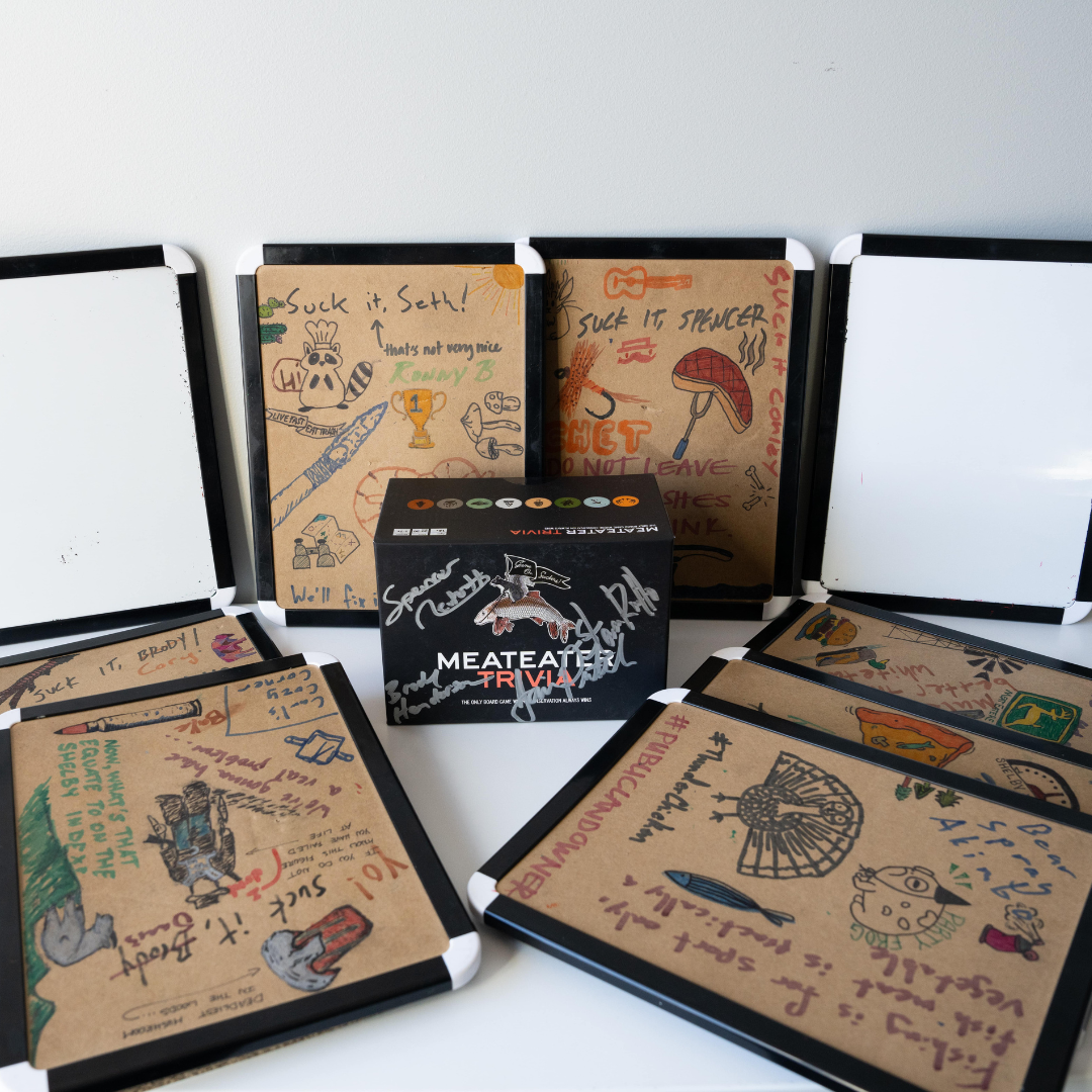 Signed black MeatEater Trivia board game box surrounded by doodle-covered whiteboards.