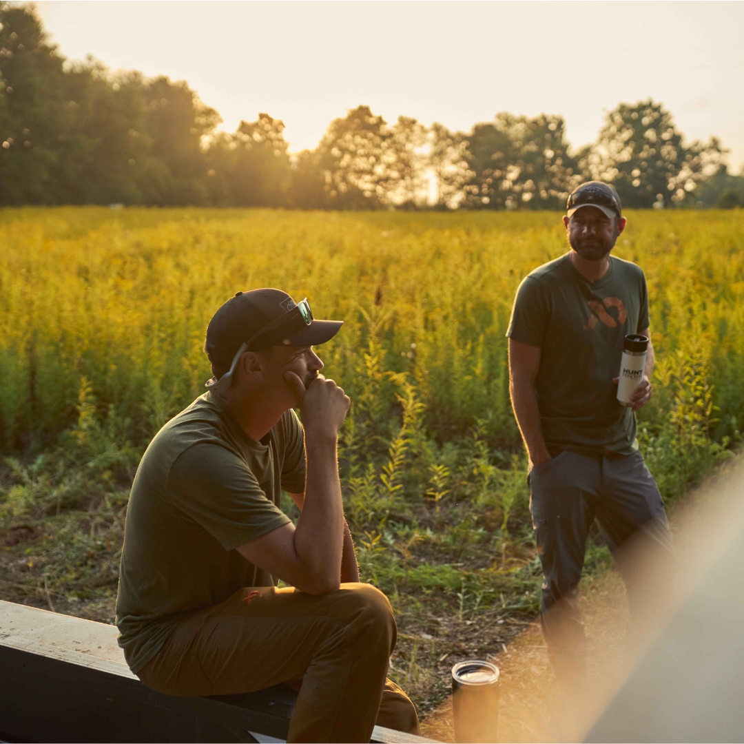 Two men wear green t-shirts, caps, outdoor pants; one holds a white tumbler in a field.