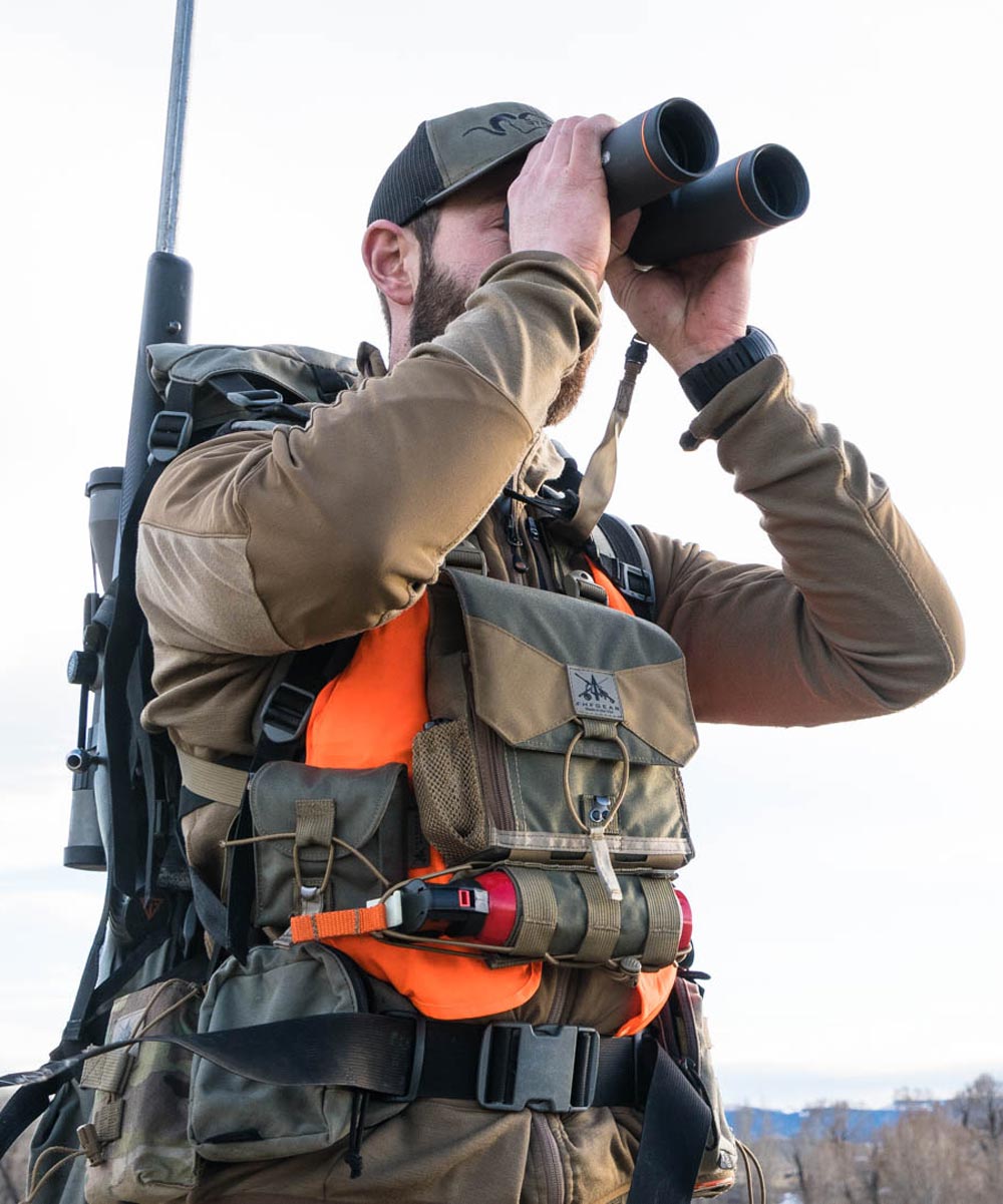 Man wears an olive green bino chest pack over orange vest, using binoculars.
