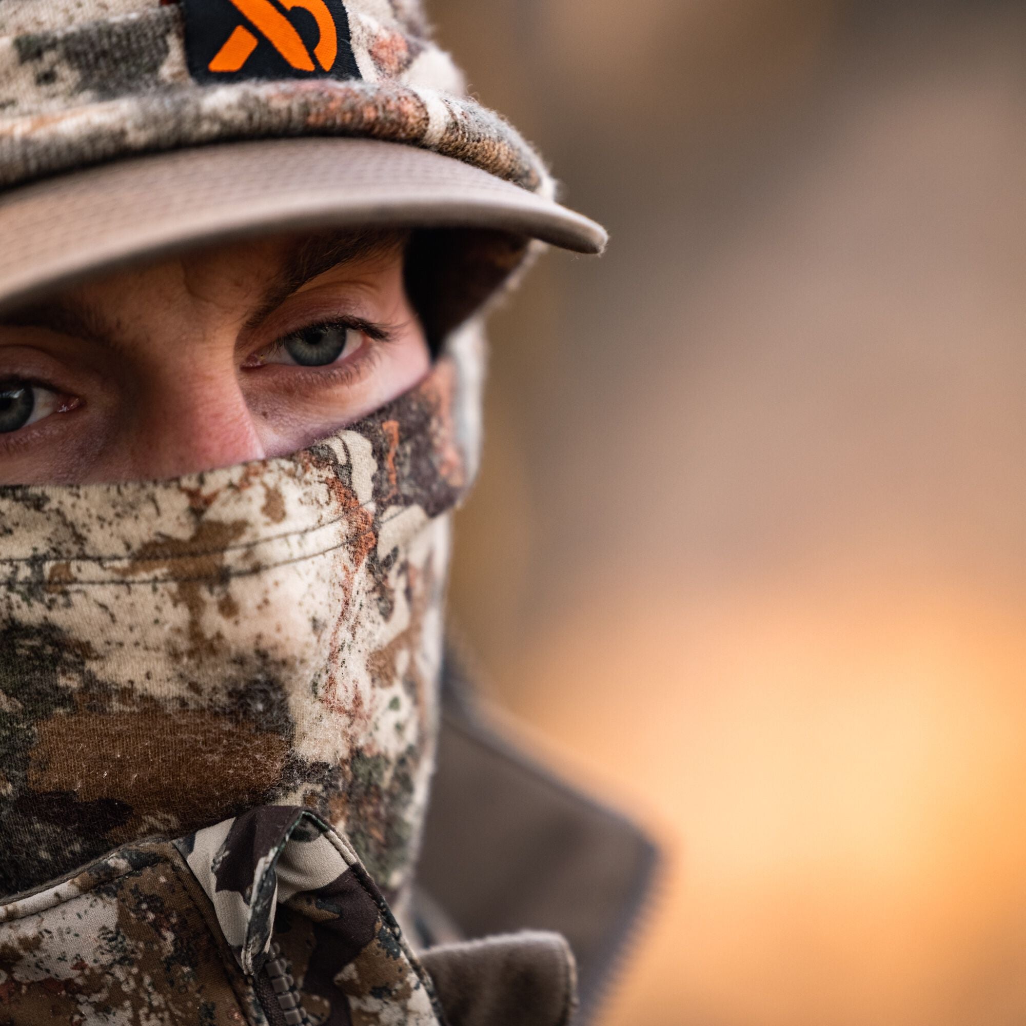 Person wears camouflage balaclava, cap with orange logo, and hunting jacket.