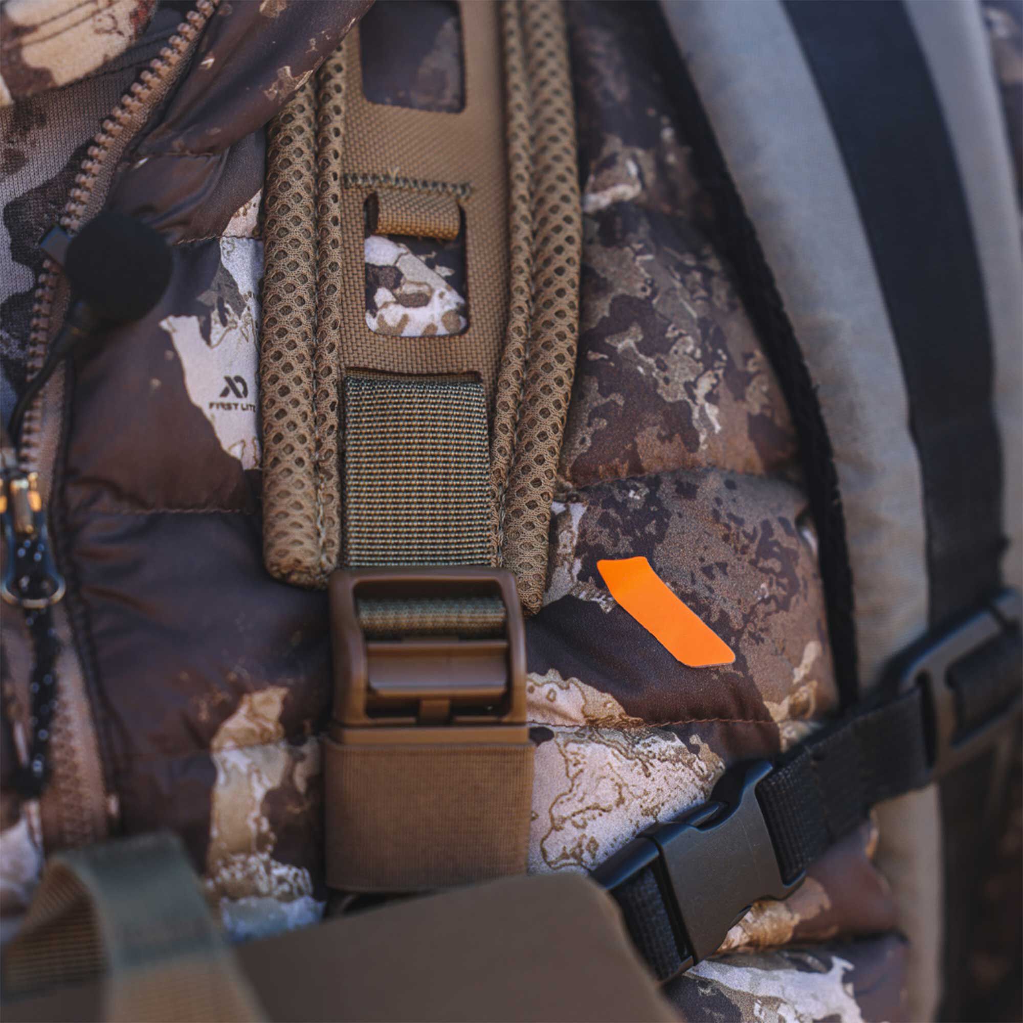 Padded camo First Lite hunting gear with tan mesh, buckles, and orange tag.