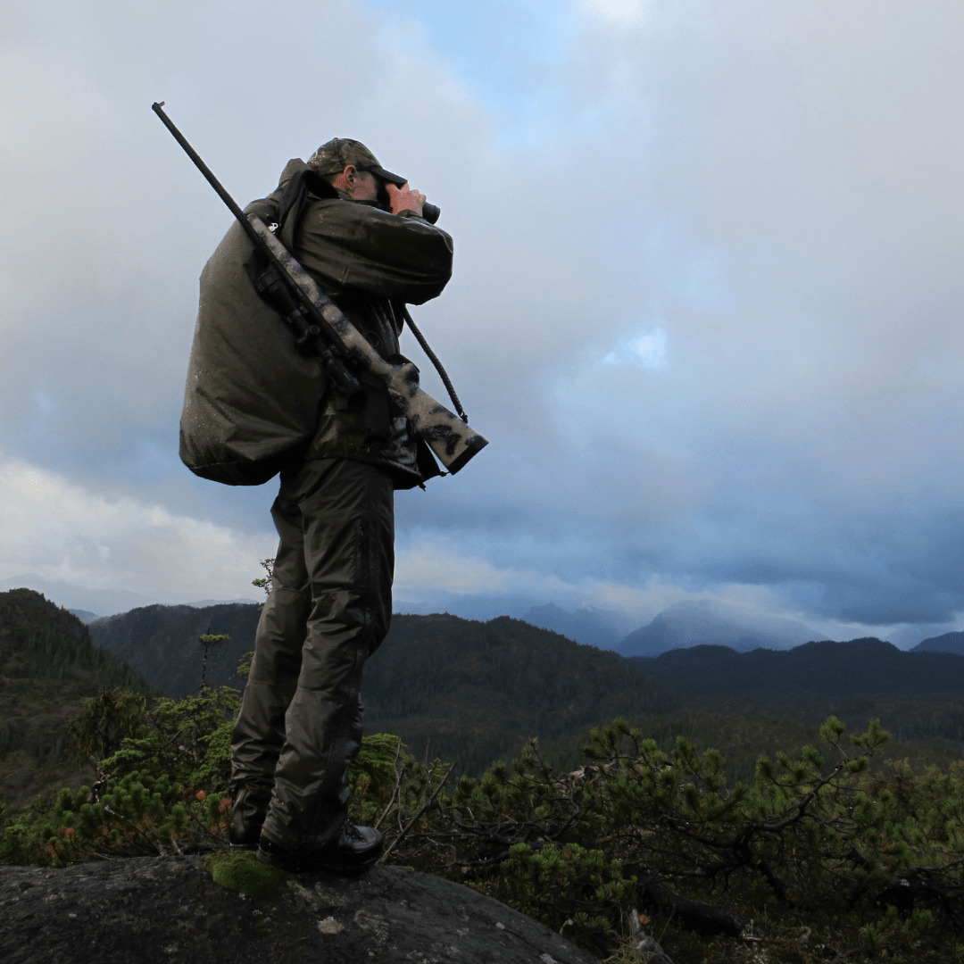 Hunter in dark green gear, carrying camo rifle & binoculars, scouting mountain landscape.