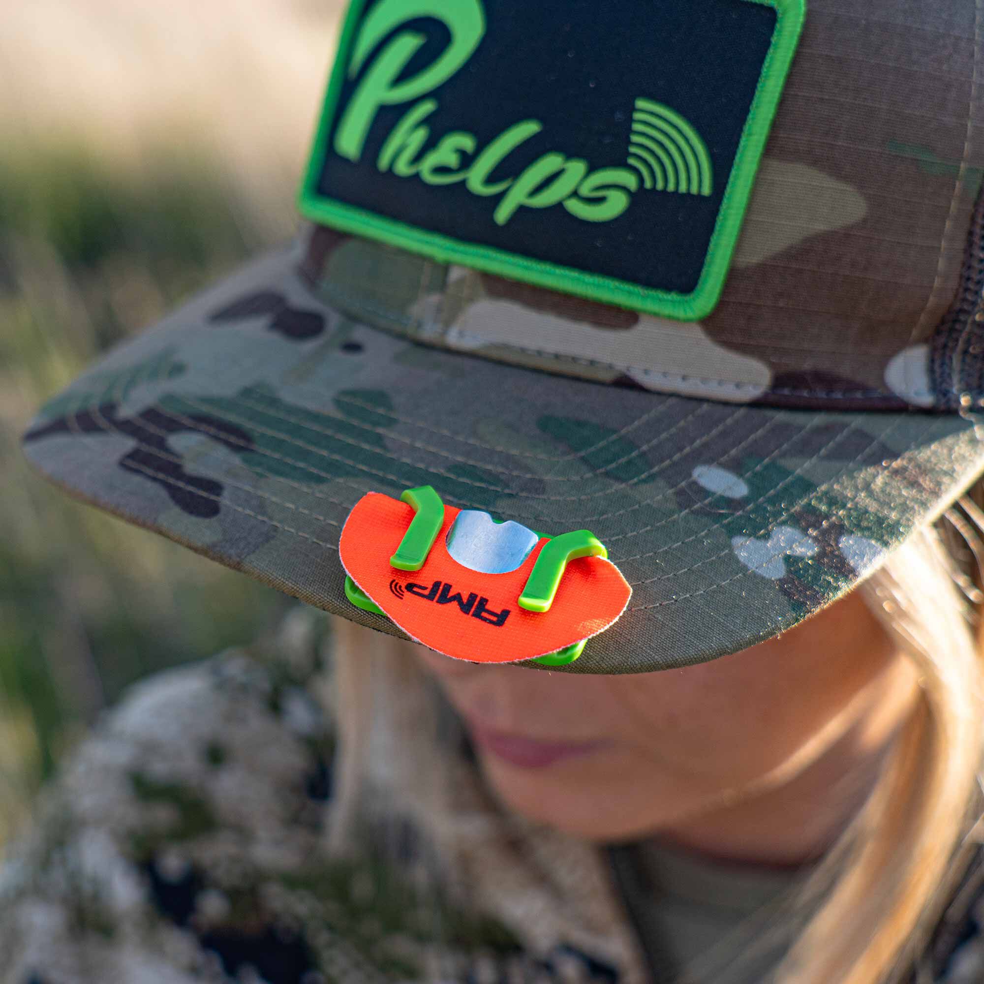 Orange AMP call clip on a camo hat, held by green grips.