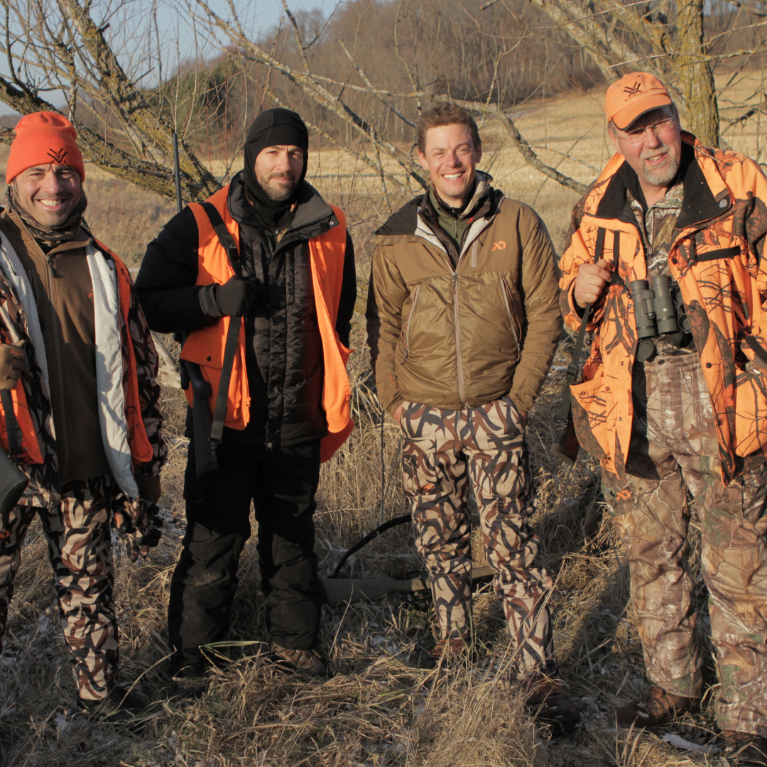 Four hunters in blaze orange vests and camo pants, one holding binoculars.