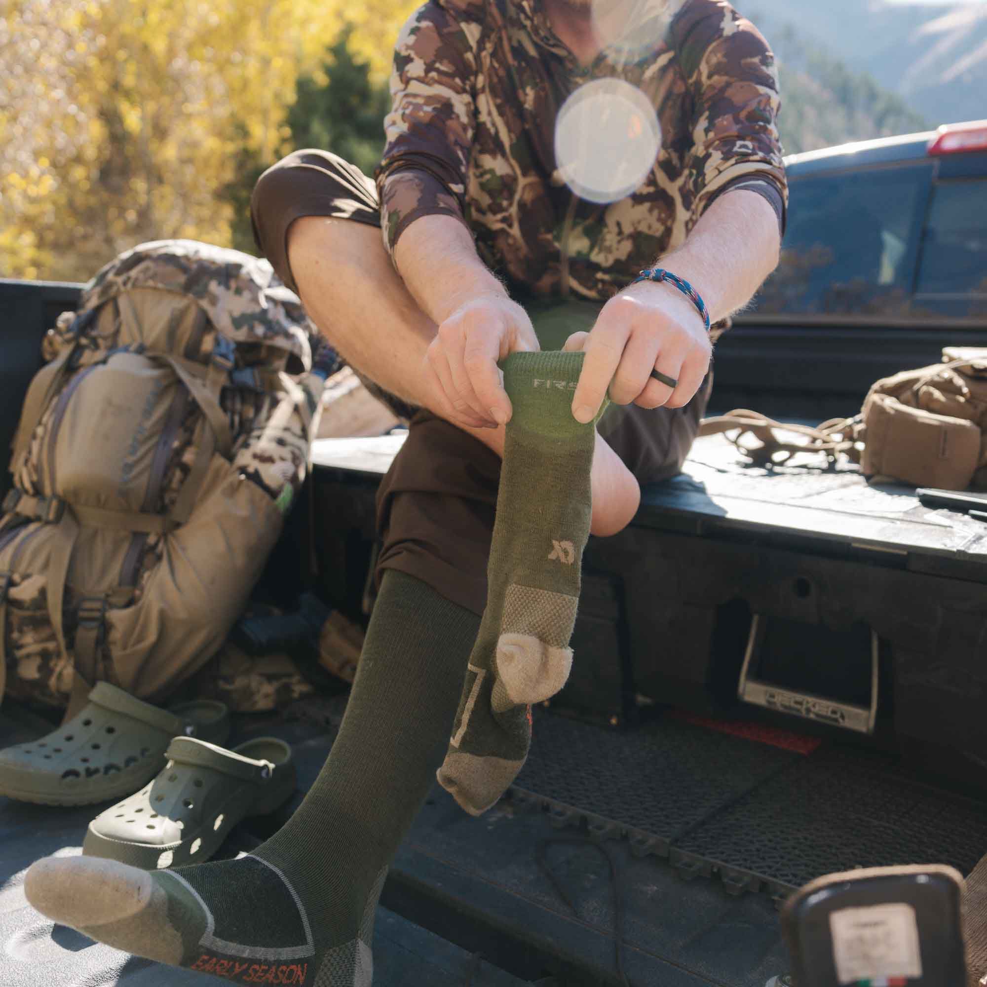 Olive green performance socks with 'FIRST' on the cuff and 'EARLY SEASON' on the heel, one being pulled on.