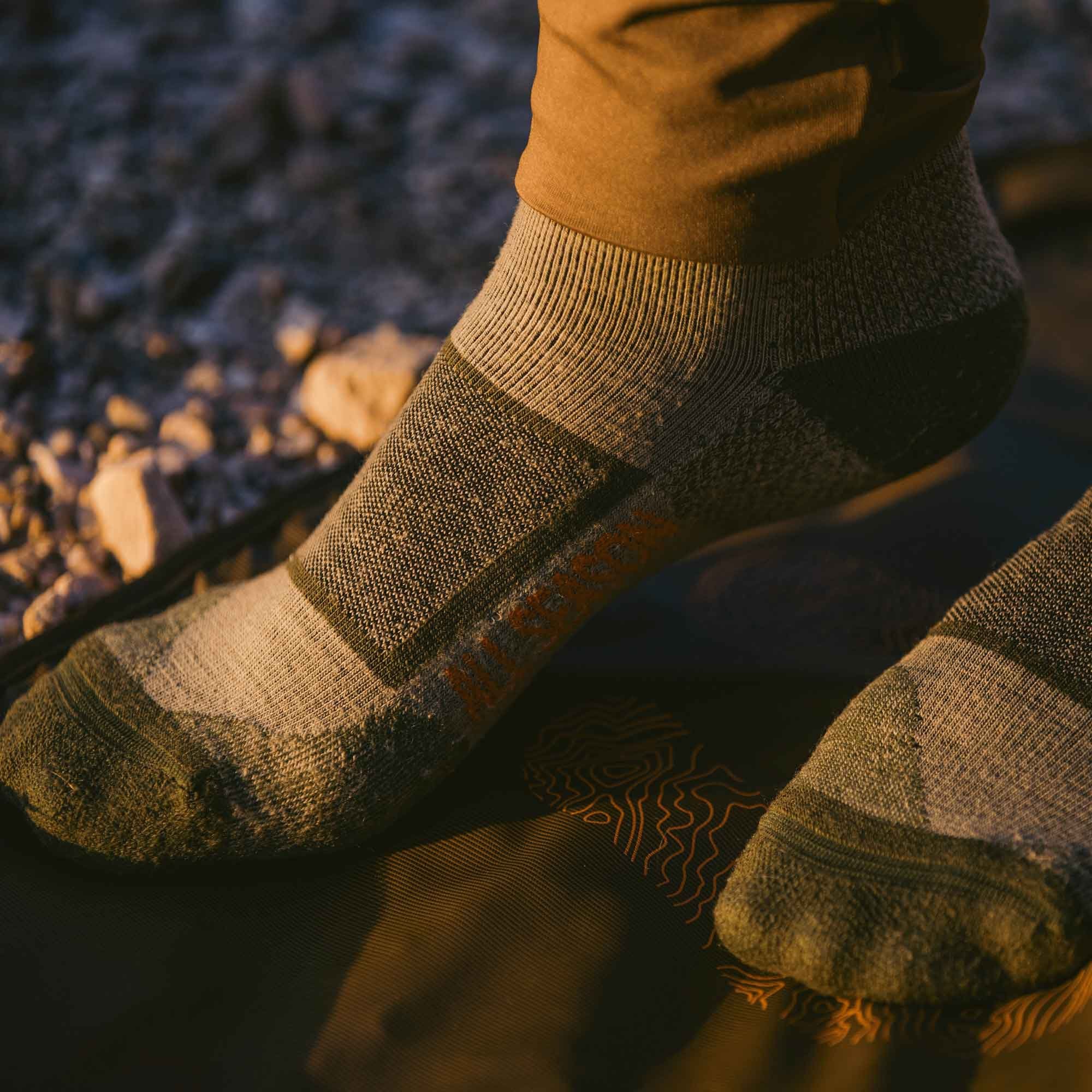 A foot wears a grey and olive green ankle sock with a ribbed cuff, textured fabric, and orange "ALL-TERRAIN" branding, resting on a dark mat.