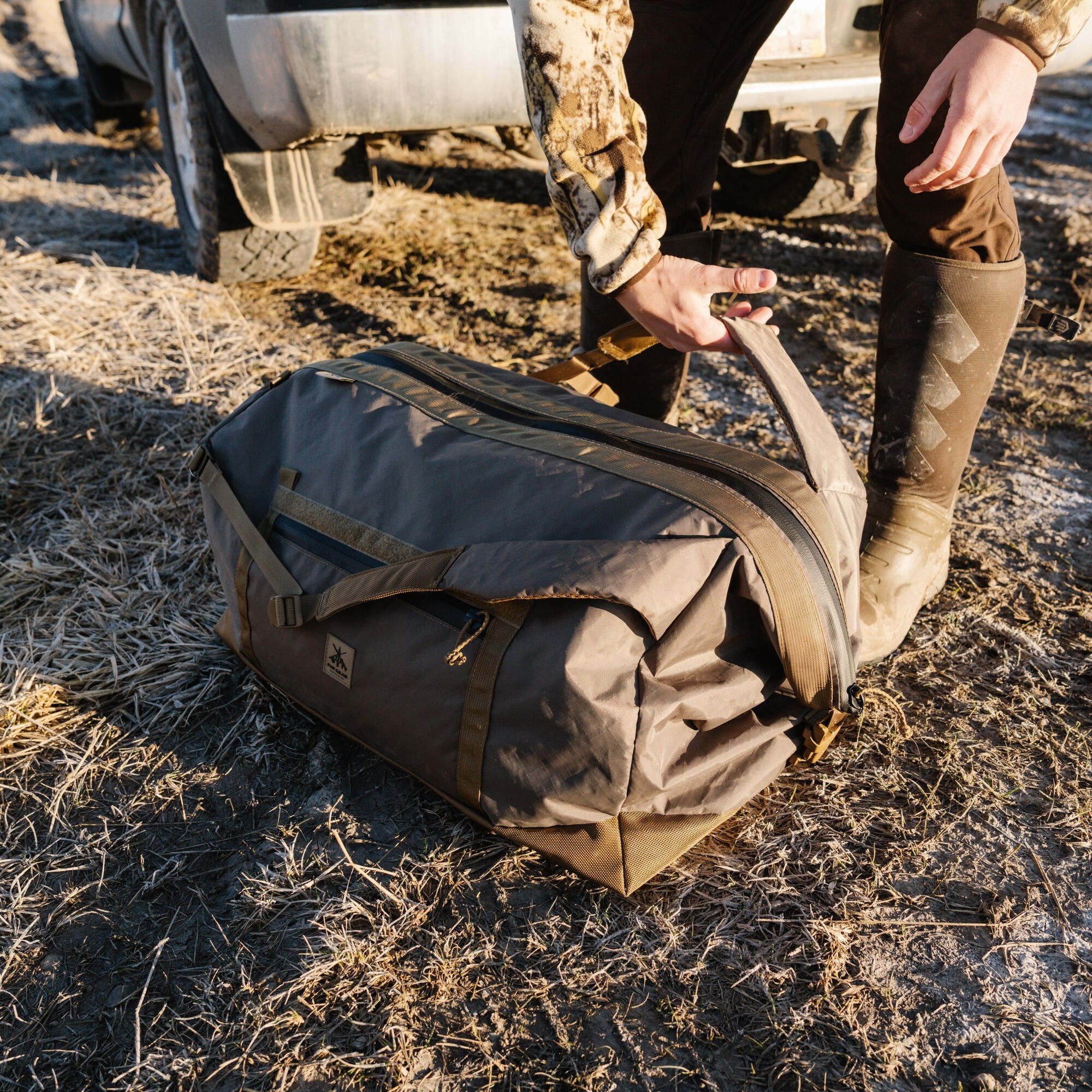 Person holding a dark olive duffel bag with tan straps, textured bottom, and logo.