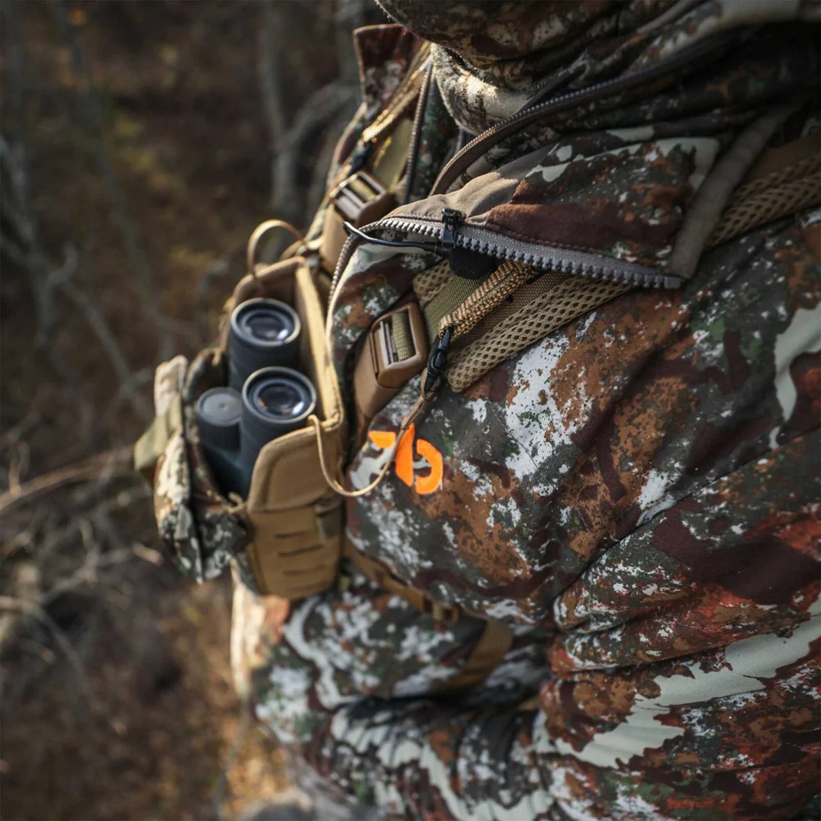 Camo jacket and tan bino harness holding binoculars worn by person; orange logo.