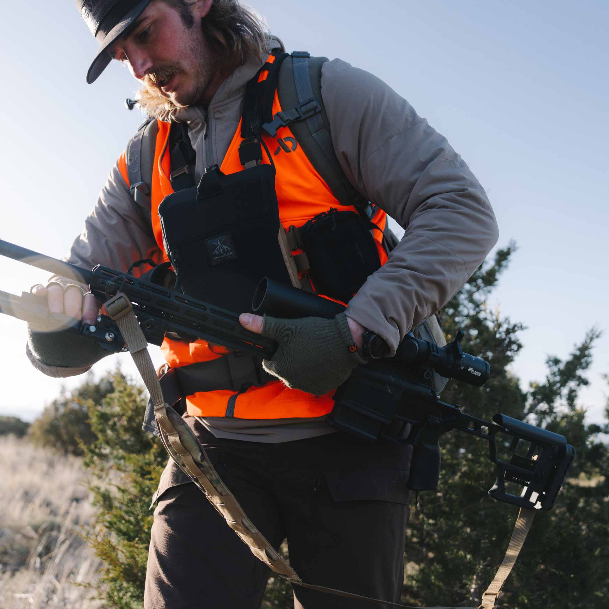Orange hunting vest with black chest rig, holding a rifle with scope & fingerless gloves.