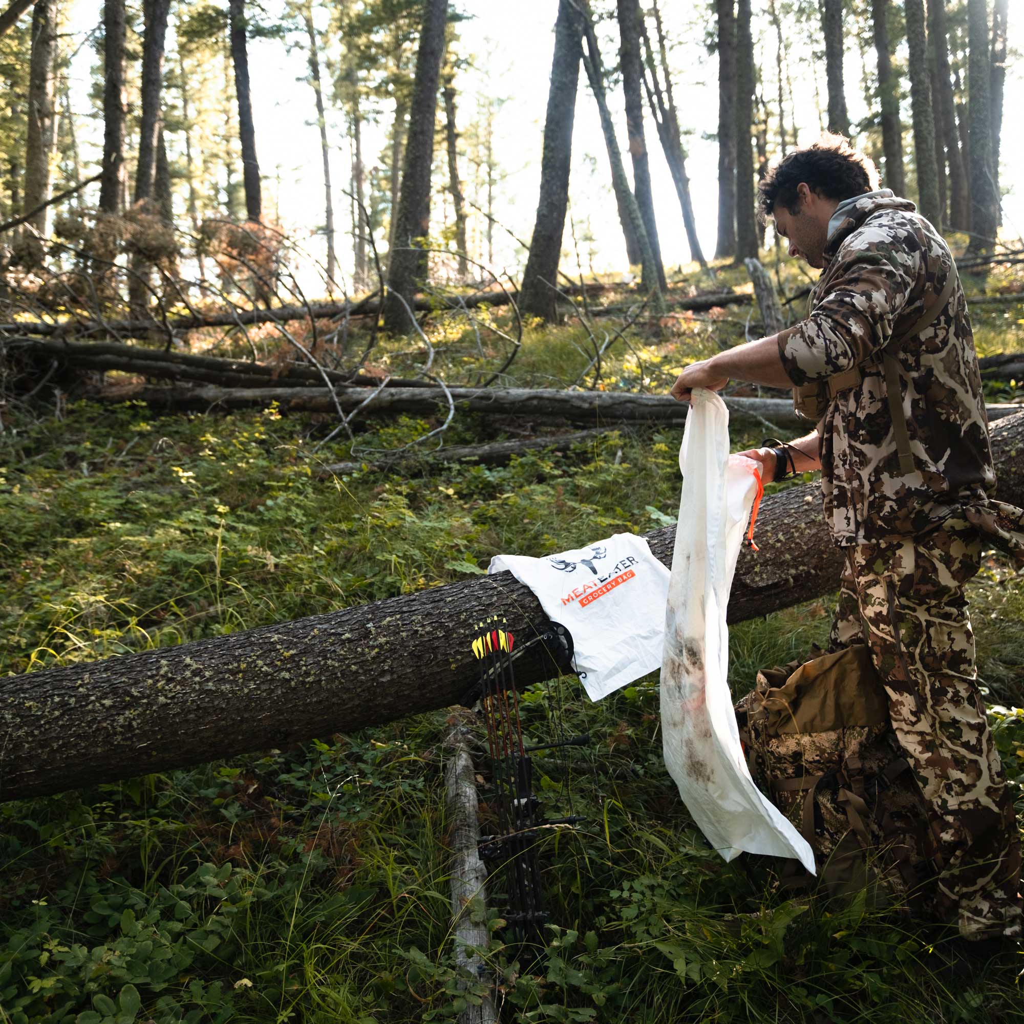 Man with a bow and wearing camouflage clothing furling open a game bag in the woods