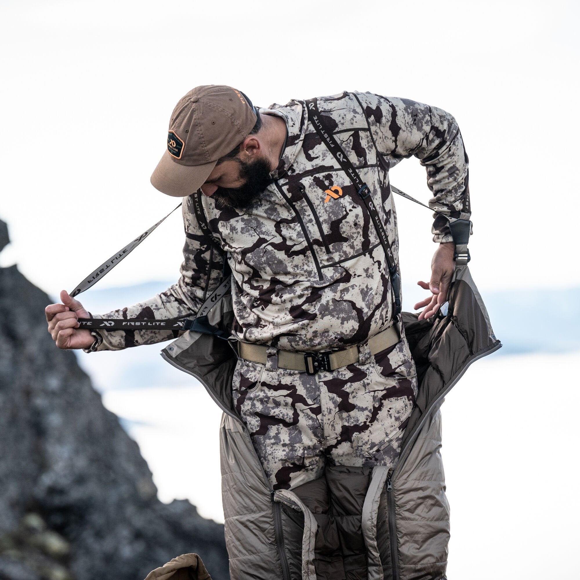 A man in a brown cap and First Lite camouflage top and pants with a beige belt, pulls up dark olive green insulated puffy pants featuring full-length side zippers and First Lite branded suspenders.