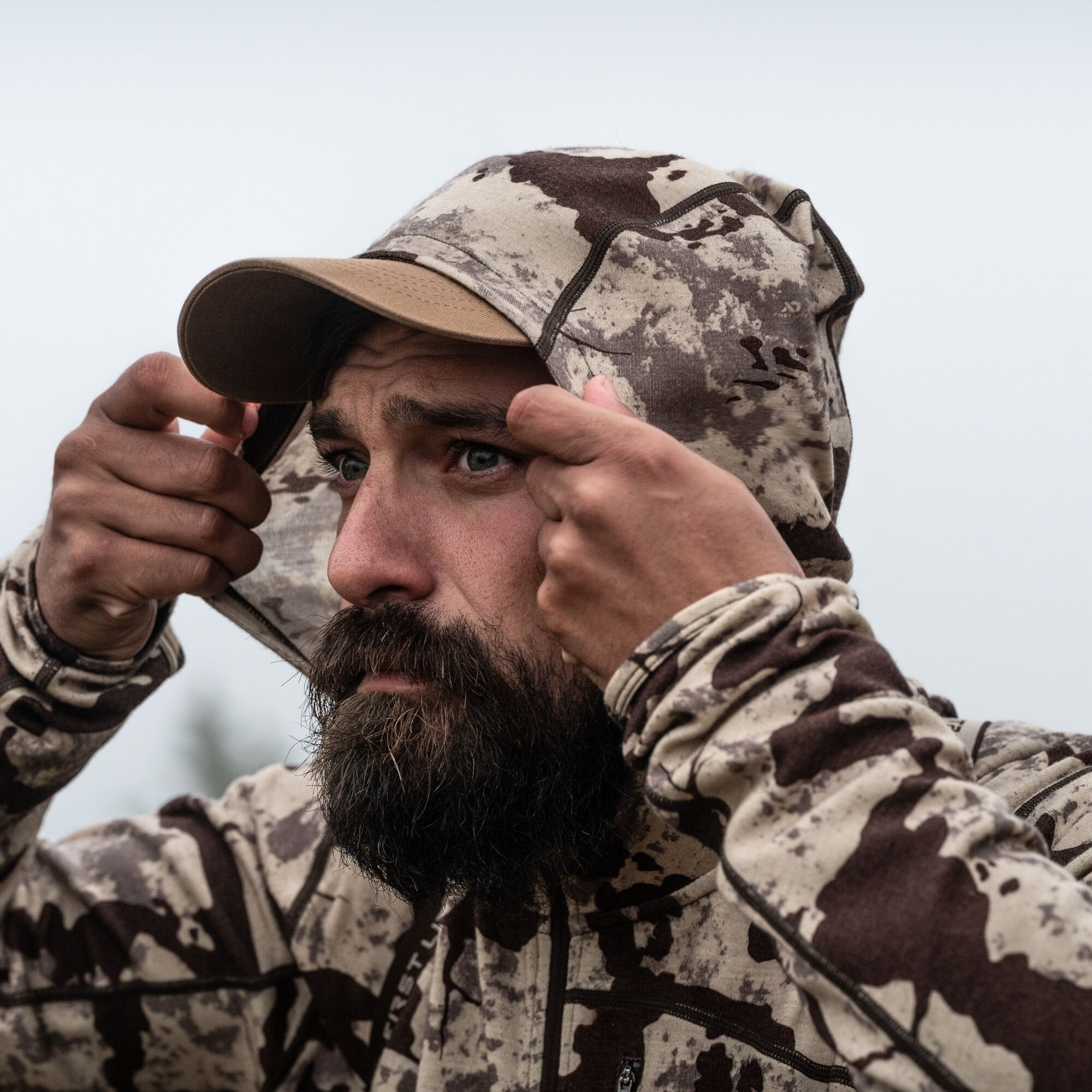 A man wearing a tan and brown camouflage hooded jacket, pulling the hood over a tan cap.