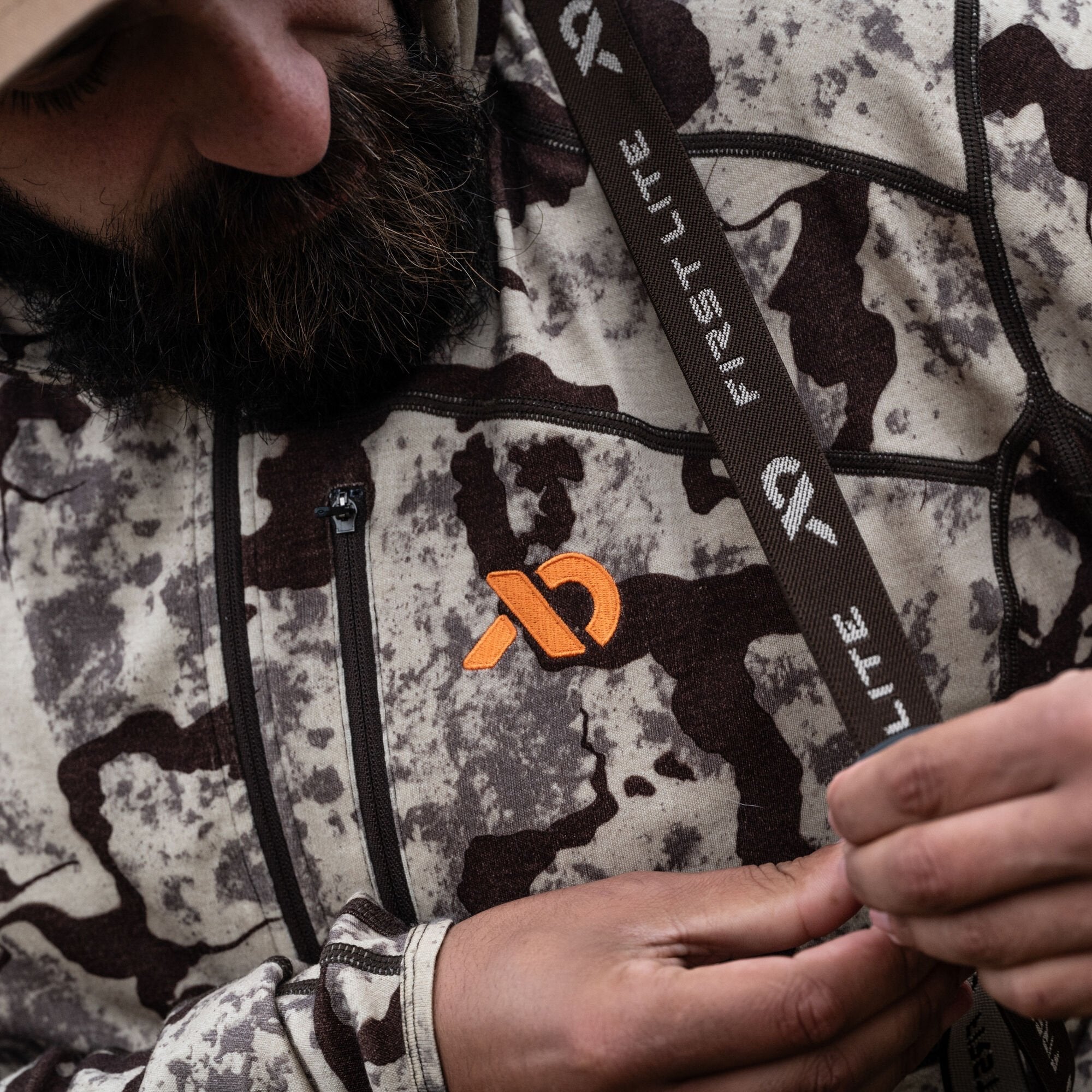 A person wears a light gray and dark brown camouflage hoodie featuring an orange First Lite logo on the chest, a zippered pocket, and a dark brown branded strap.
