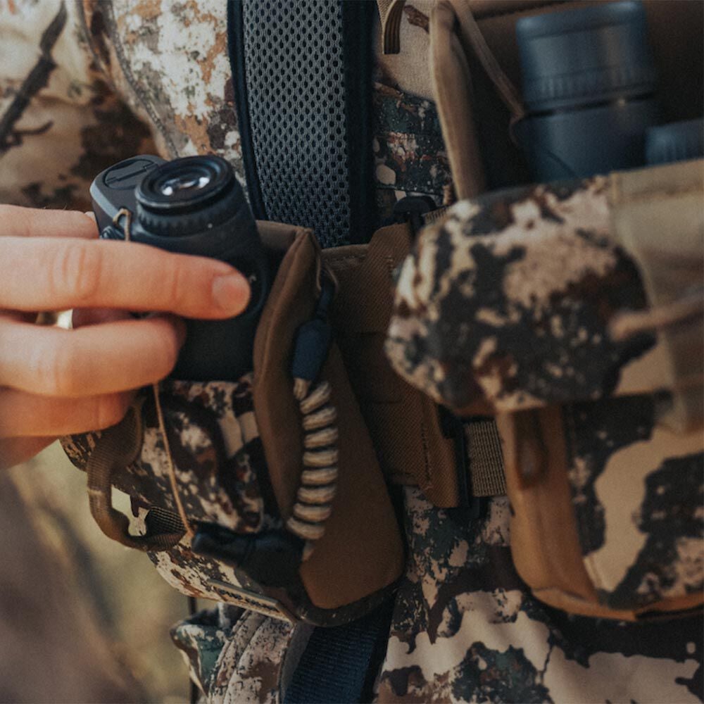 Hand places black rangefinder into tan pouch on a camo chest rig with coiled lanyard.