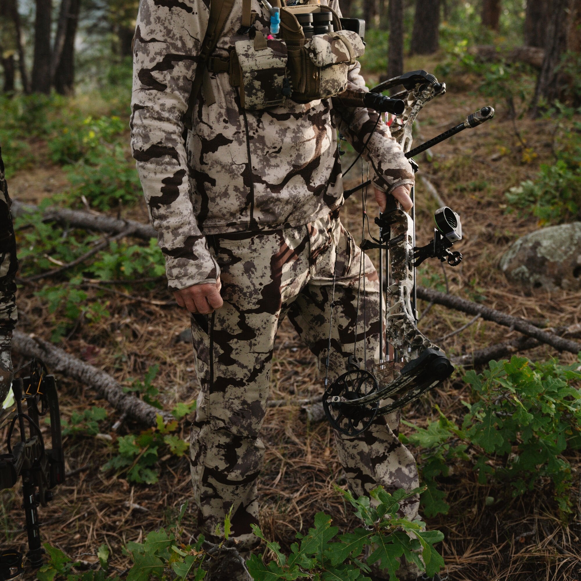 A hunter wears a full light tan and dark brown blotchy camouflage hunting suit with a chest rig, holding a matching compound bow in a wooded environment.