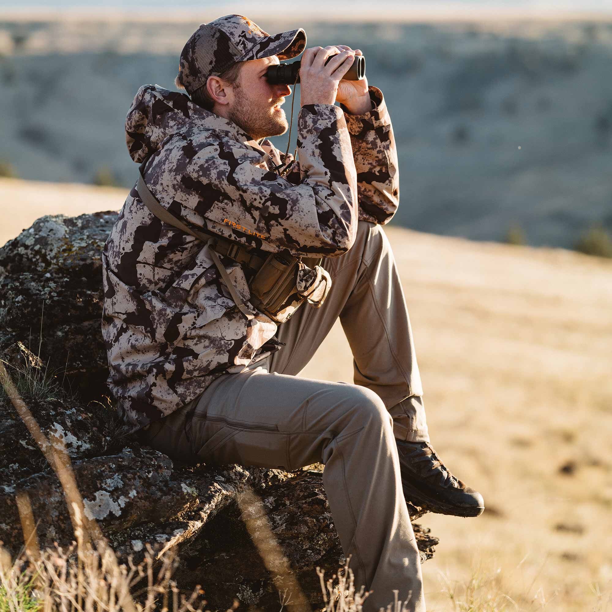 Man wearing a First Lite Fusion camo hooded jacket, matching hat, and tan pants while looking through binoculars.