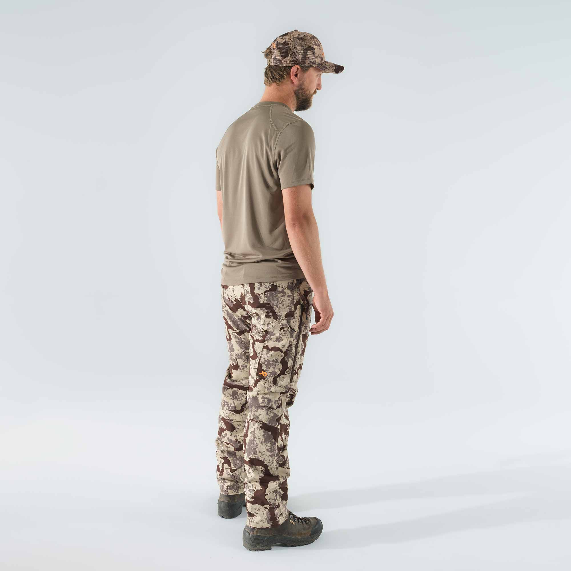 Man wearing a taupe short-sleeve shirt, brown and beige camouflage cargo pants with side zippered pockets and an orange logo, and a matching camouflage cap.