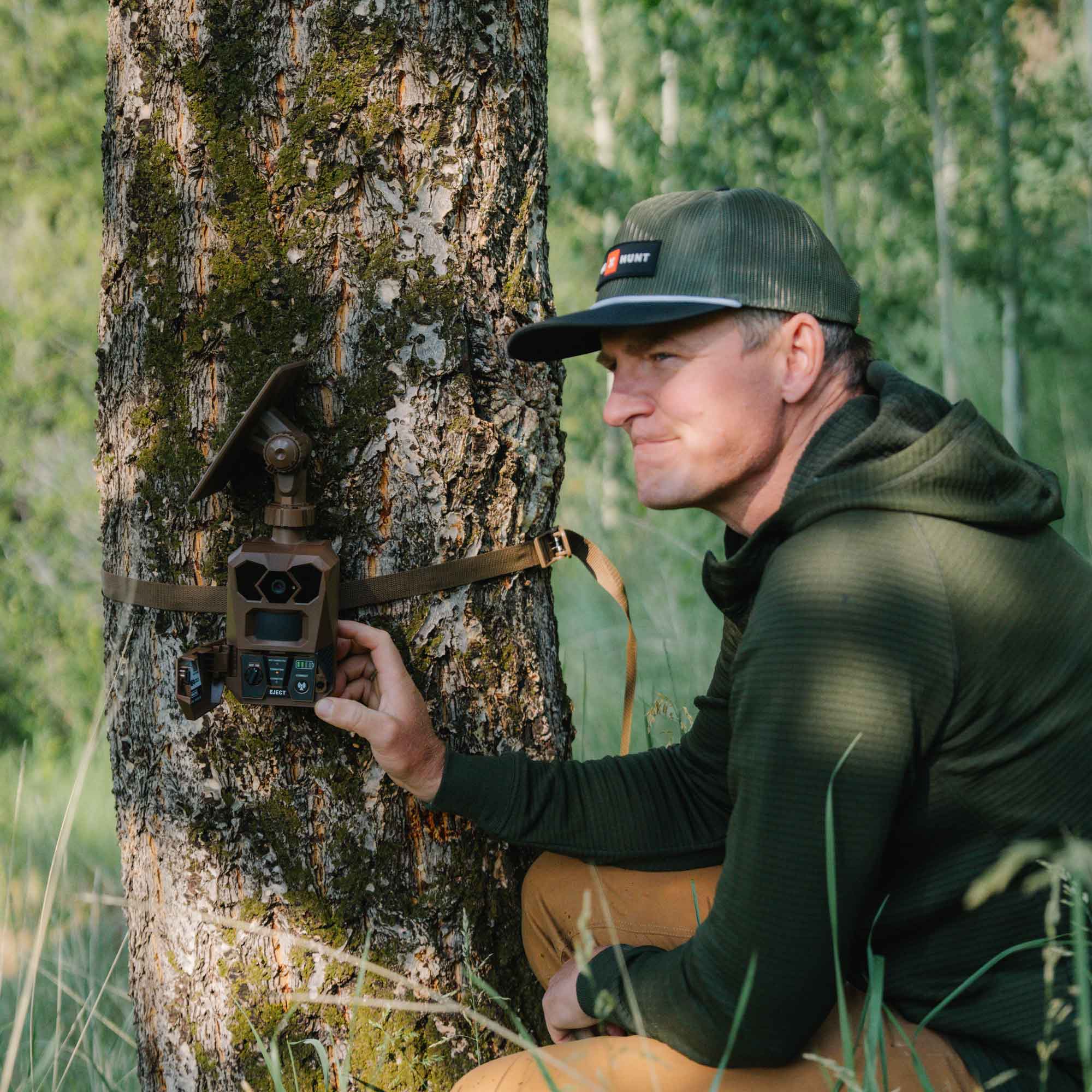 A man in a forest next to a tree, adjusting a brown trail camera mounted on the trunk with a strap.
