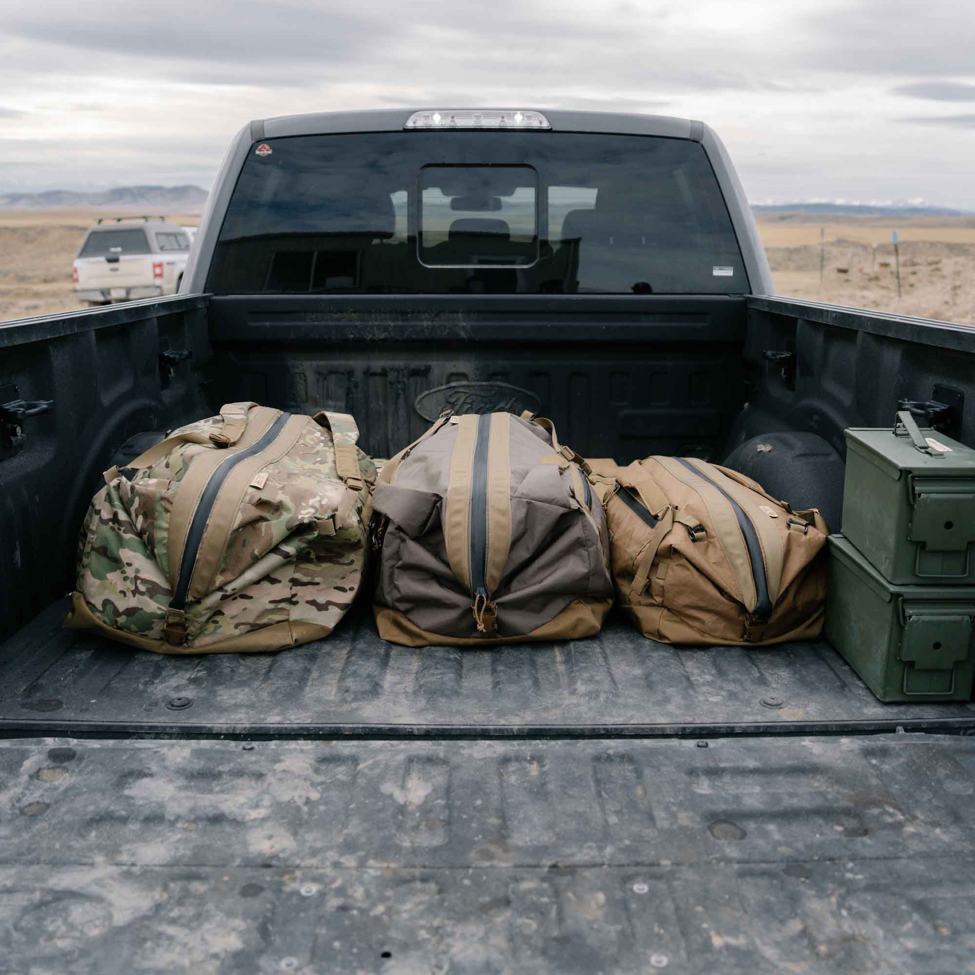 Three duffel bags: camo, brown, and khaki, with central zippers in a truck bed.