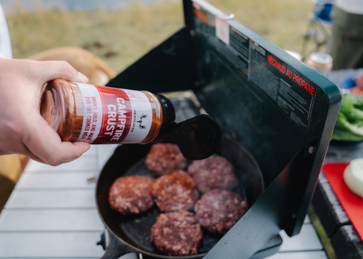 Person seasoning burgers on a grill with 'Campfire Crust' seasoning.