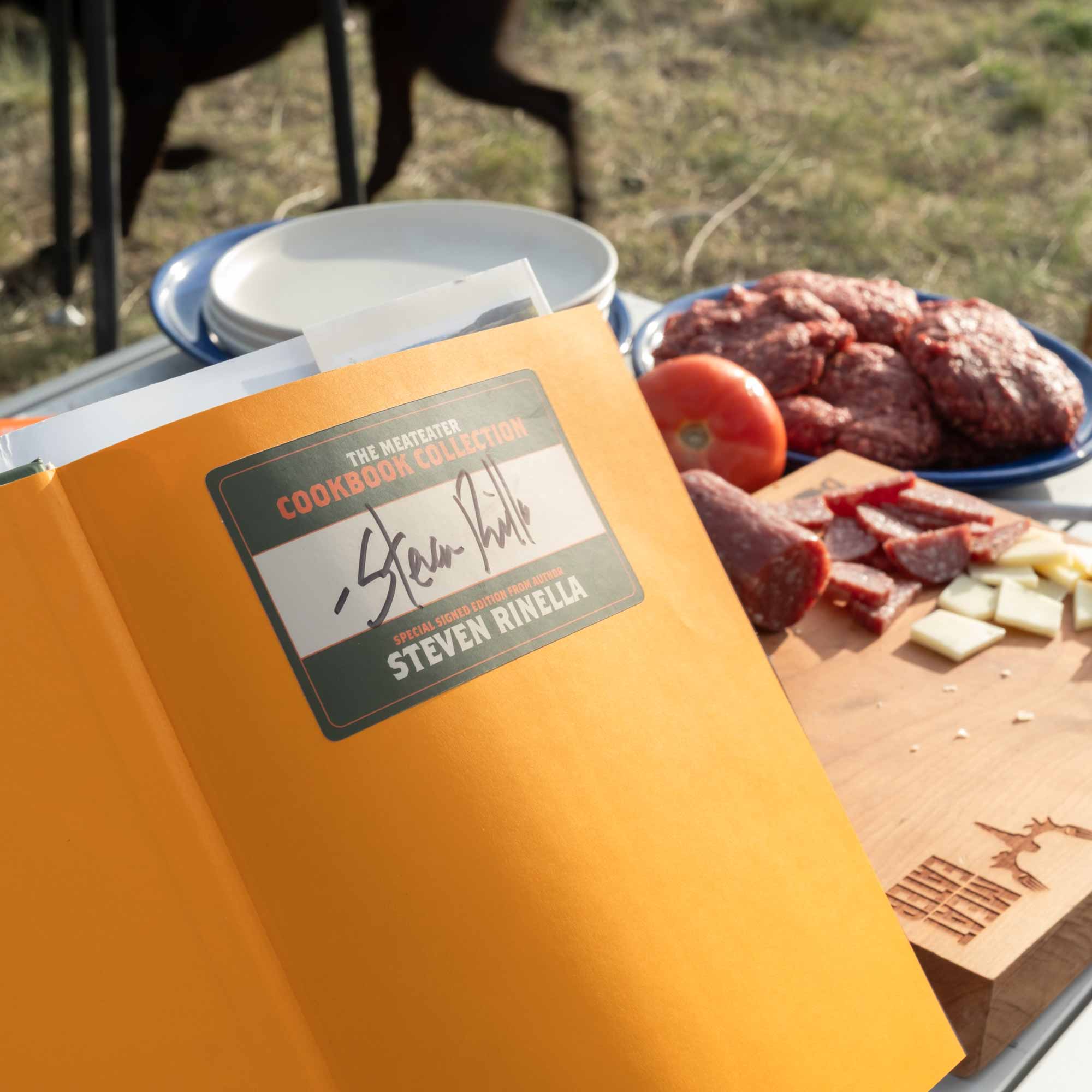 Cookbook with a signature on the cover next to a cutting board with meats and vegetables outdoors.