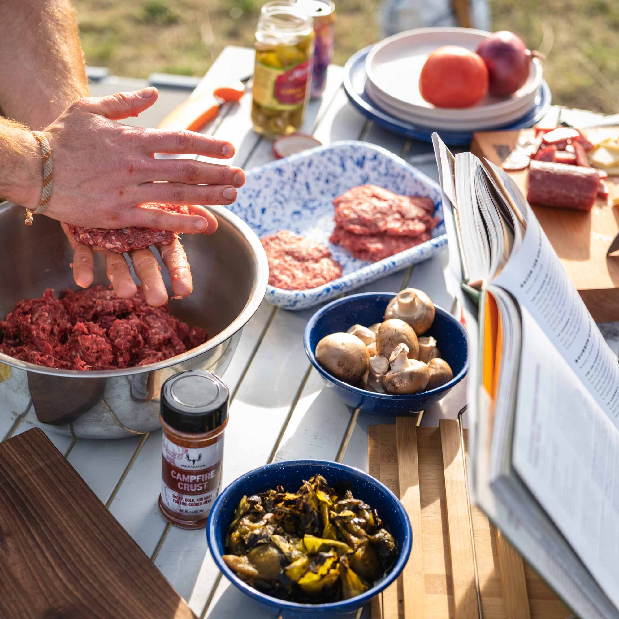 Person preparing food outdoors with ingredients and a recipe book on a table.